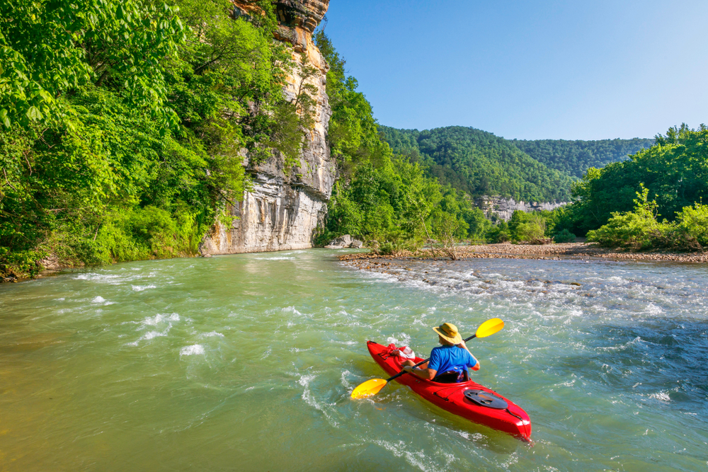 red kayak with man on it gently paddling along the river with small rapids one of the bucket list of things to do in arkansas 