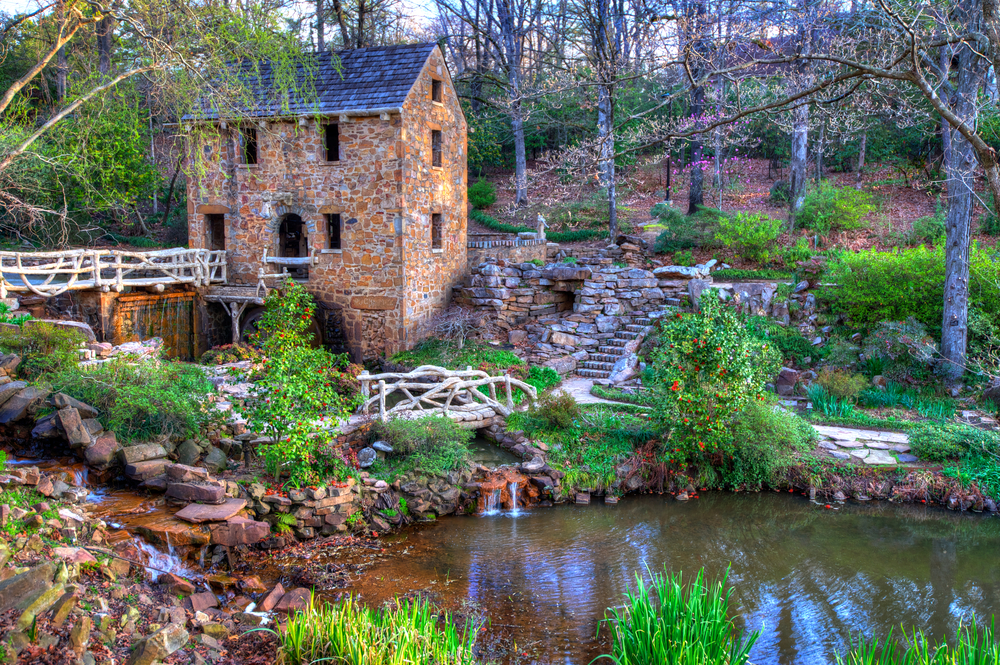 The Old Mill in brown and red stone standing over small river with green hedges and brown bridges around it