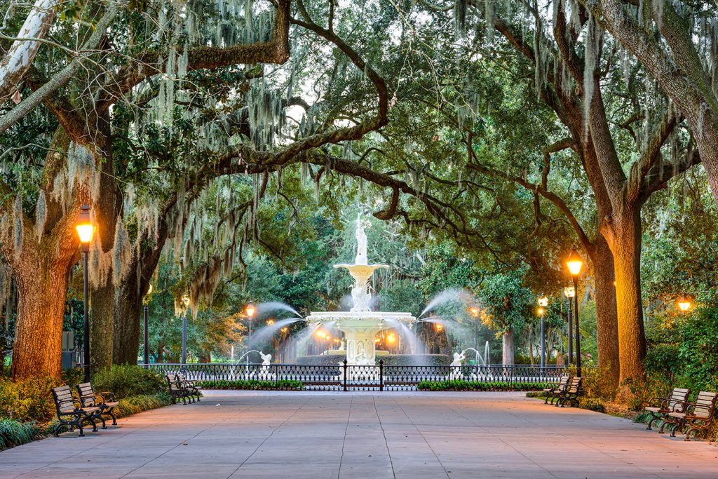 Moss-covered trees hang over street lamps, benches, and a running fountain during an evening at Forsyth Park, visiting which is one of the best things to do in Savannah.