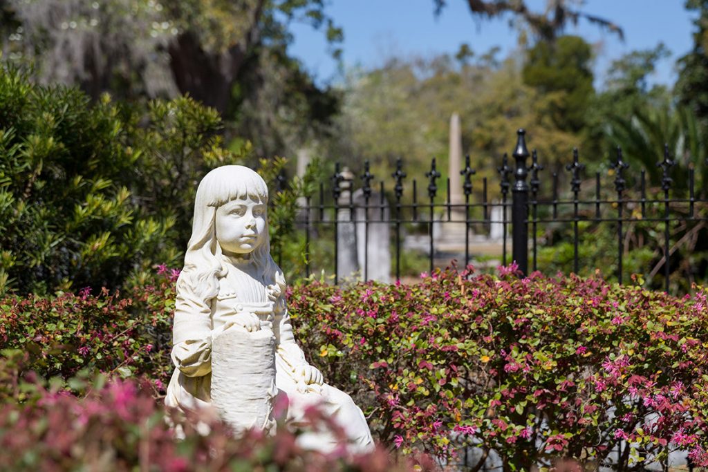 The marble statue of a young girl, known as the Gracie Watson Statue, surrounded by a flowering bush and other graves at in Bonaventure Cemetery in Savannah. 