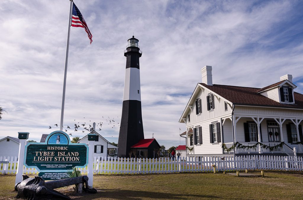 The black and white striped Tybee Island Lighthouse, with surrounding white picket fencing and buildings, and a flock of birds flying past.