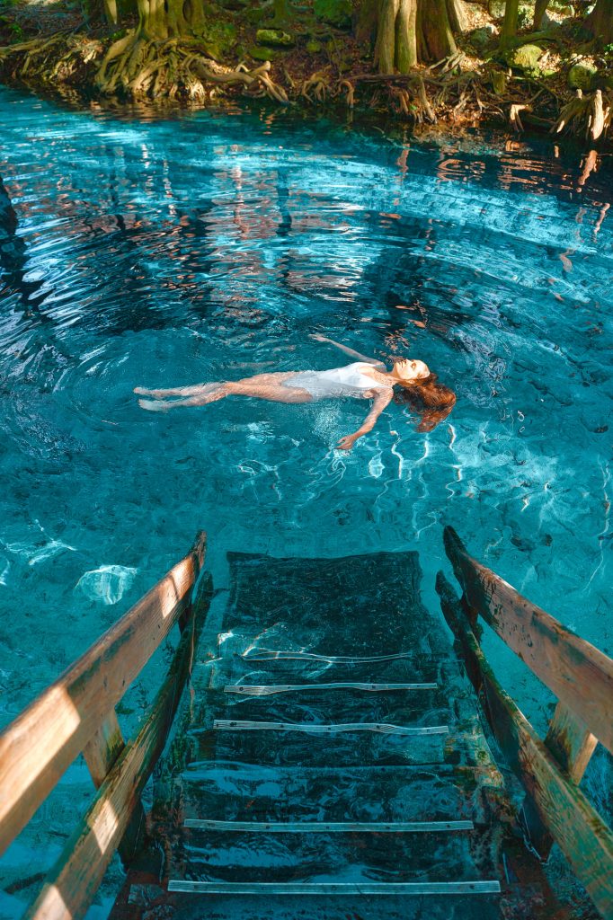 woman in white swimsuit and red hair laying in blue water with stairs leading into it