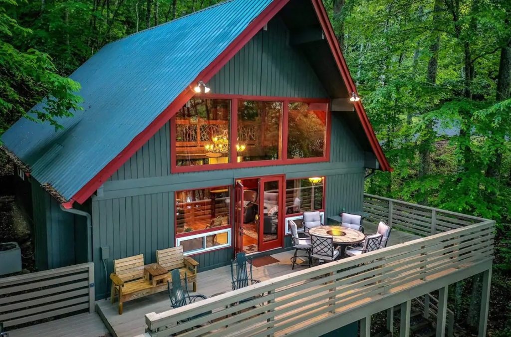 A slightly overhead view of the front of a large A-frame cabin with a large front porch surrounded by woods