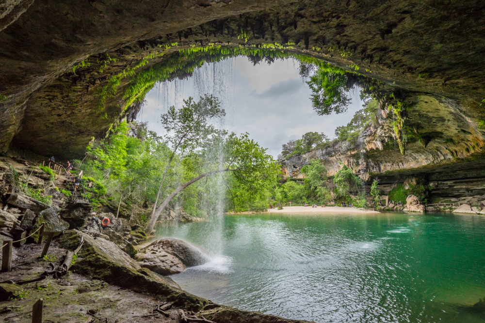 The scenic view of a small waterfall cascading over the mouth of a cave into a grotto filled with aqua water makes Dripping Springs one of the most refreshing waterfalls in texas