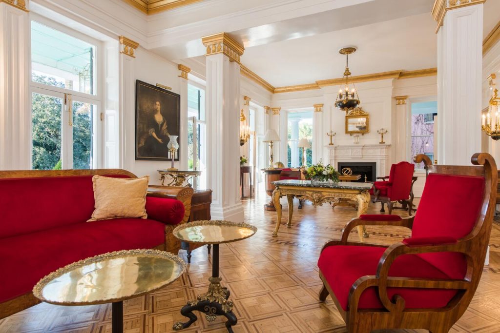 The lobby of an ornately decorated bed and breakfast. There are white walls, gold trim, ruby red furnishings, and gold tables and chandeliers. There are windows on every wall. 