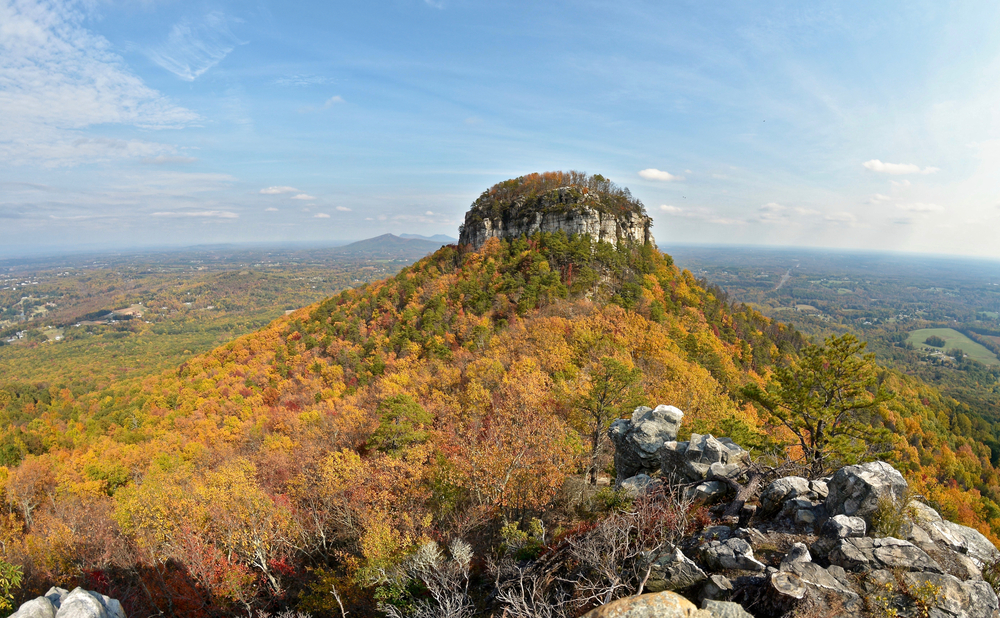 A photo of Pilot Mountain in Pilot Mountain State Park covered in orange, yellow, and green trees of fall in North Carolina.