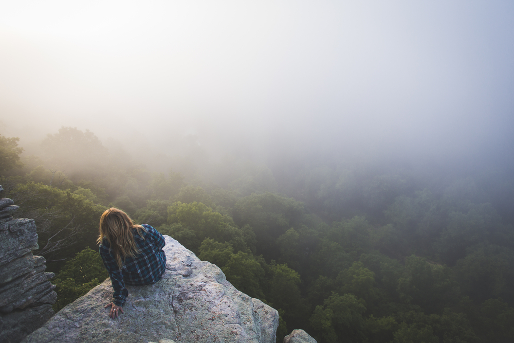 A photo of a girl sitting on a rock cliff overlooking a foggy forest in Arkansas.
