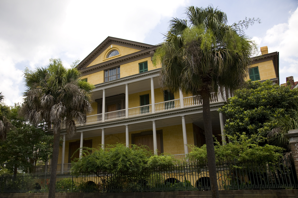 The exterior of the Aiken-Rhett House. The house is yellow with a large front porch and a large front deck. The house is surrounded by palm trees and shrubs. 