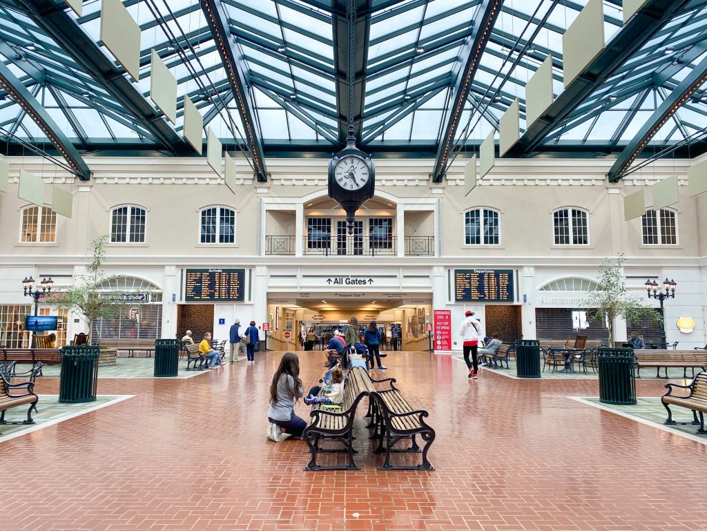 The lobby of the small airport in Savannah Georgia. It has wrought iron and wood slat benches, a glass ceiling, and an antique style clock hanging from the ceiling. The walls are white and cream and made to look like a small village. There are people walking to a hallway leading to "All Gates" as well as people sitting on the benches. Big screens on the wall show departures and arrivals.