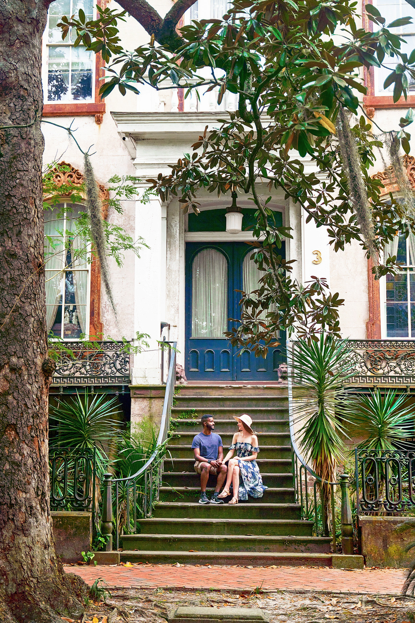 A couple sitting on the stone steps in front of the Alex Raskin Antique store in Savannah Georgia. The steps are surrounded by greenery and lead to double doors with large windows that are painted blue. The building is old and painted white with rust colored trim. The woman in the couple is wearing a blue floral sundress with a sunhat. The man in the couple is wearing a blue shirt and khaki shorts. They are facing each other on the steps and holding hands. A haunted and cool building you should see during your 3 days in Savannah