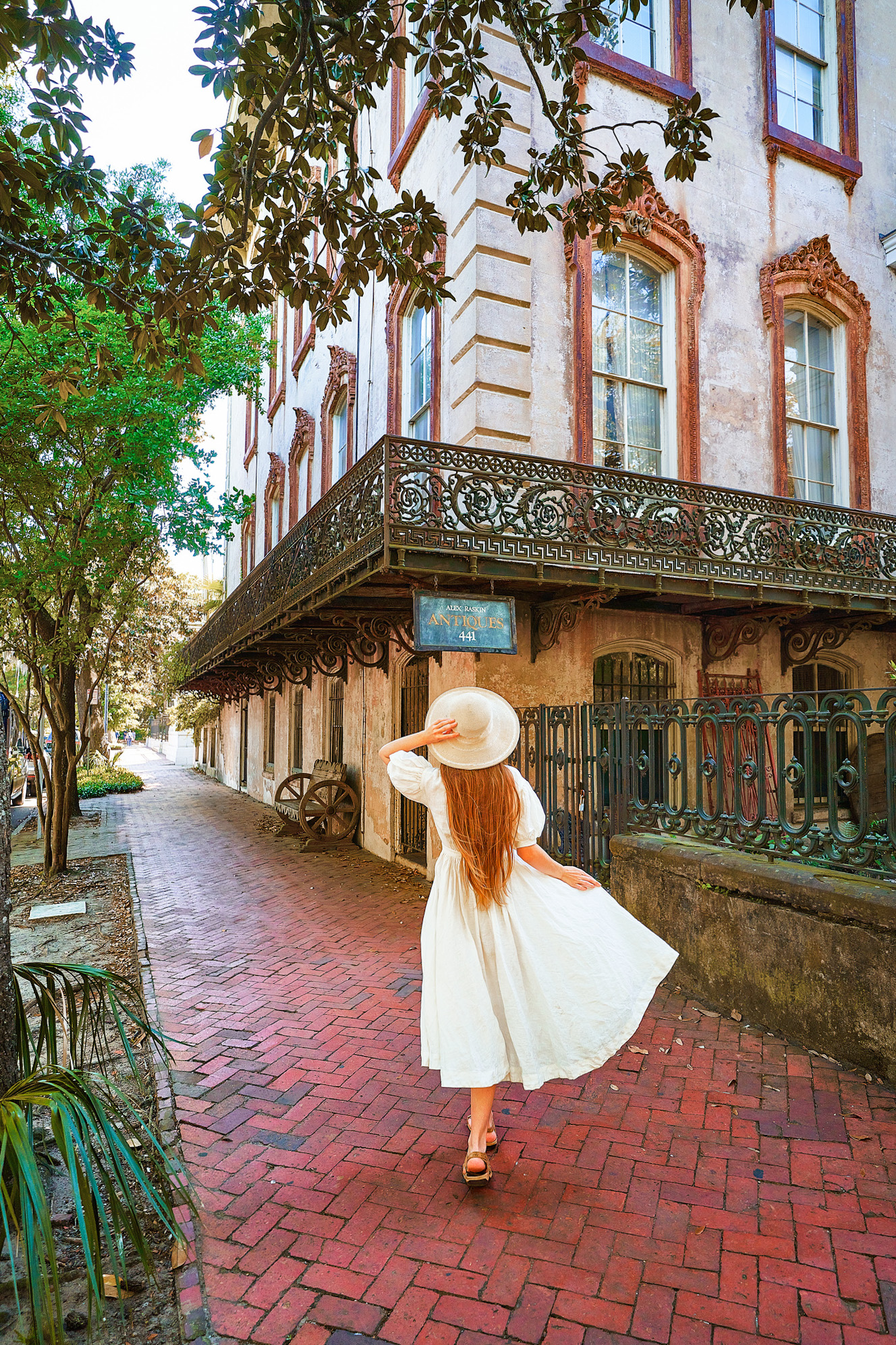 A woman in a white sundress with long hair and a white sunhat standing outside of a run down mansion. She is standing on a brick sidewalk looking up at the building. The building has stained plaster, rusted window trimmings, and a rusted wrought iron balcony and gate.
