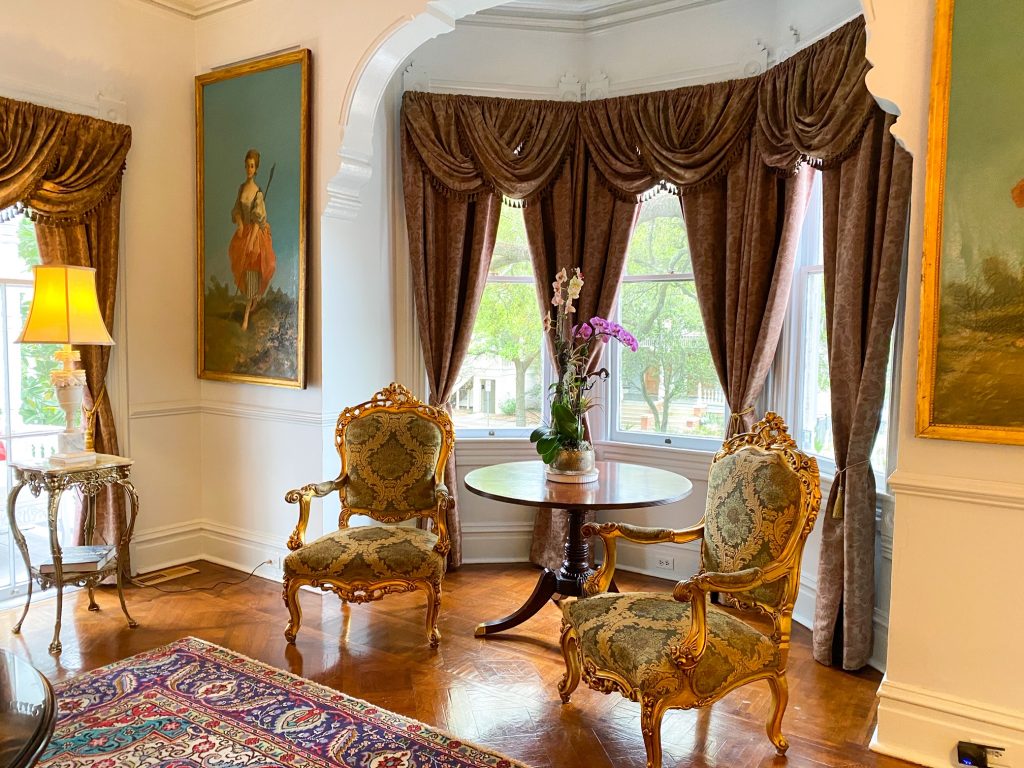 The opulent interior inside the Amethyst Garden Inn in historic Savannah. There are ornate gold chairs with brocade fabric, three windows with thick drapes, and paintings on the wall. A beautiful historic inn that is a great place to stay during your 3 days in Savannah
