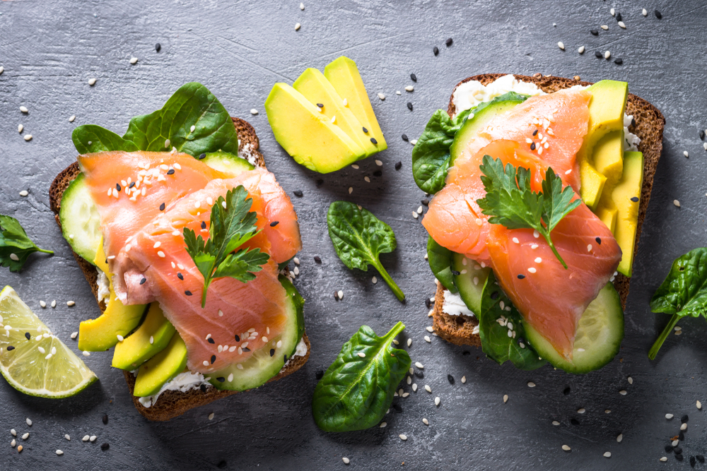 A plate of avocado toast with cucumbers, spinach, lox, and sesame seeds on it. It is on a black plate and there or lime wedges near it. 