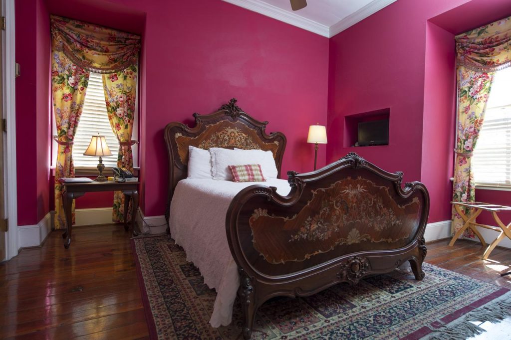 An opulently decorated bedroom with fuchsia walls, a large and ornately painted bed, and floral curtains on the two windows. There is an oriental rug under the bed, its a great place to stay during a weekend getaway in Charleston