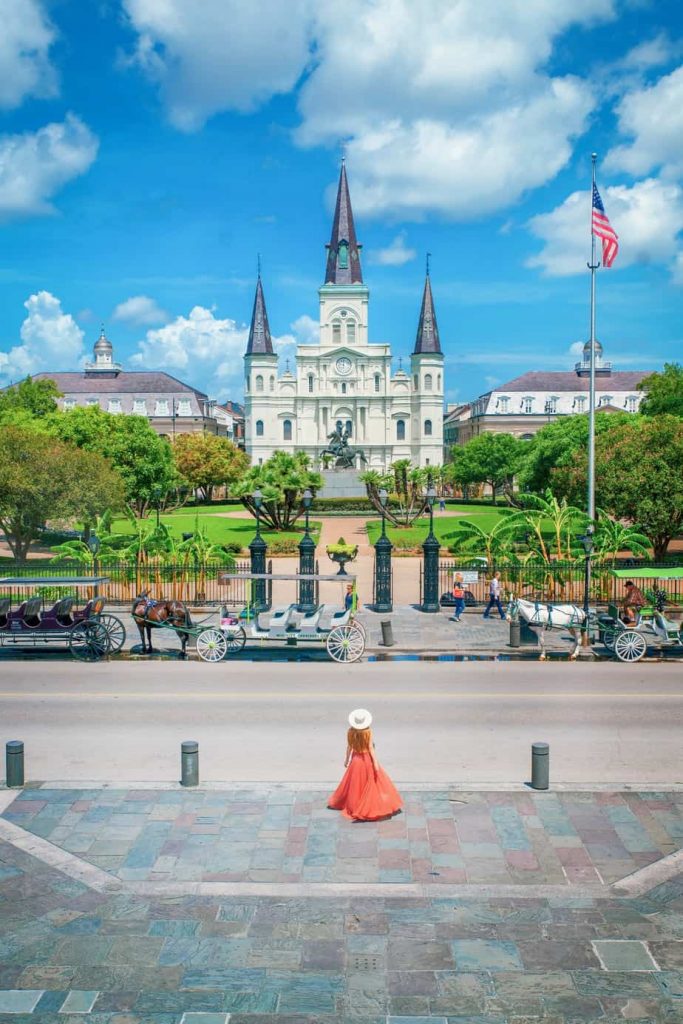 Victoria stands in an orange dress outside as horse drawn carriages wait for passengers.