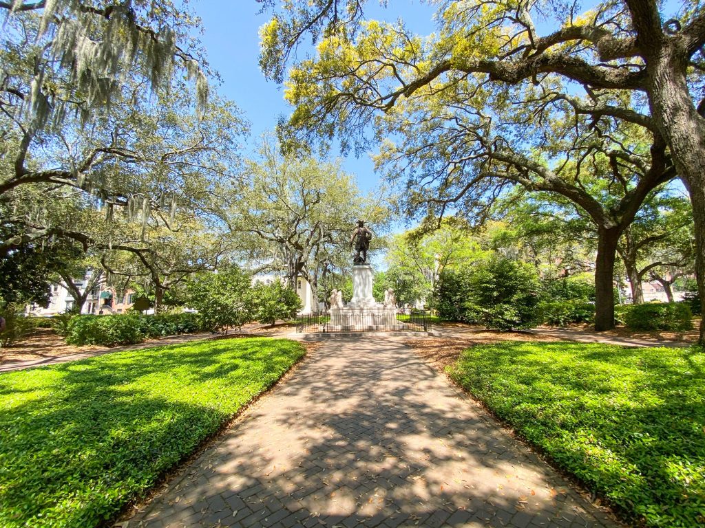 The view of one of the squares on Bull Street in Savannah.  It has sidewalks surrounded by grass leading up to a statue. The statue is a large metal statue of a man on a large white pedestal. There are a lot of trees and shrubs surrounding the square. A great place to see on your 3 days in Savannah trip