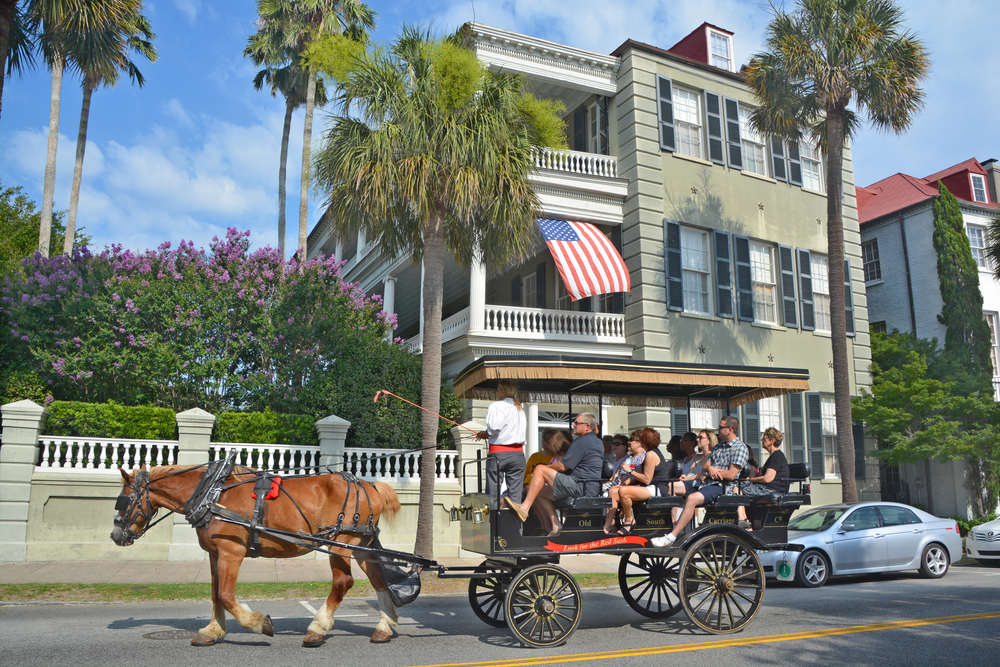 People in a carriage being driven by a horse in front of a historic house in Charleston South Carolina. The house is green and there are palm trees and a lilac bush in front of it. A great way to explore a weekend in South Carolina