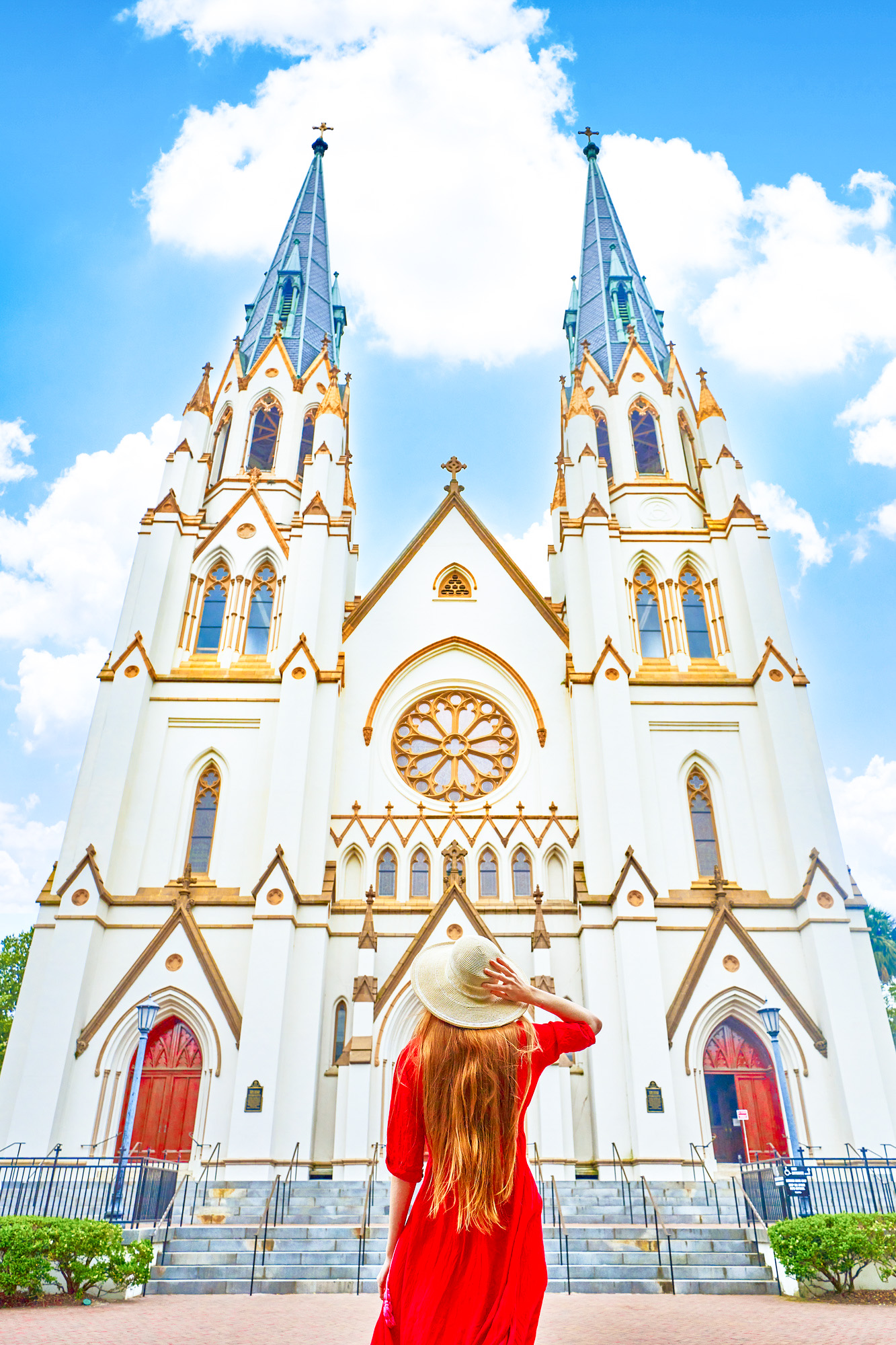 A woman standing in a bright red dress with her long hair down and wearing a cream colored sun hat looking up at the Cathedral Basilica of St. John The Baptist. The Cathedral is very large and cream with an intricate Gothic Revival architectural design. The trimmings of the church are gold and there are two large arched doors on either side of the building that are bright red. 