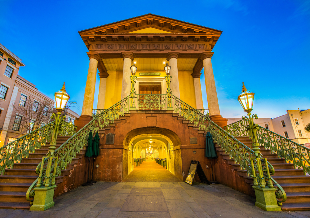 The entrance to the Charleston City Market in downtown Charleston. The entrance has two large staircases on either side leading to the main doors. It is sunset, so the building is glowing yellow and all lit up.