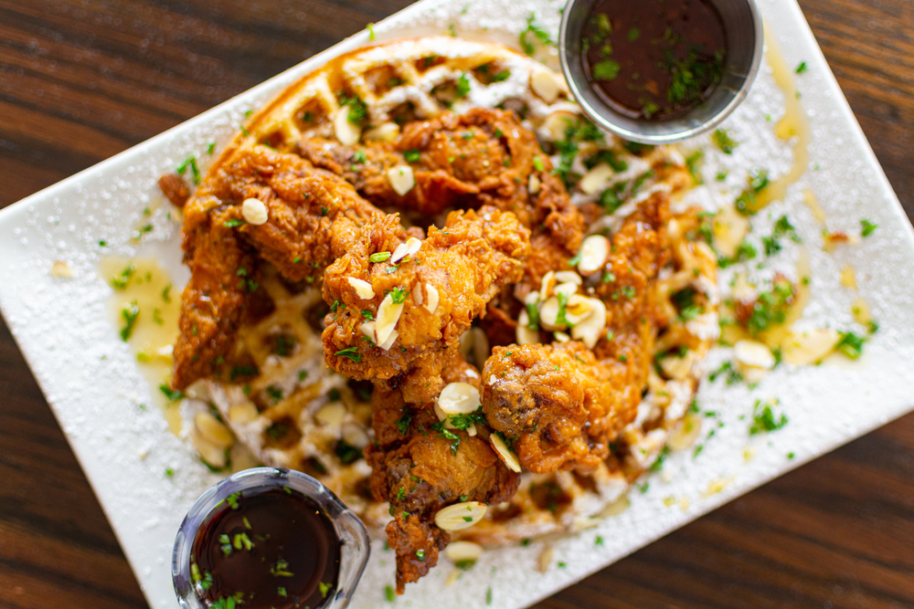 A plate of fried chicken over waffles. There are toppings on it like scallions and nuts. There are also two dipping sauces on the rectangular white plate. The plate is on a wood table. 