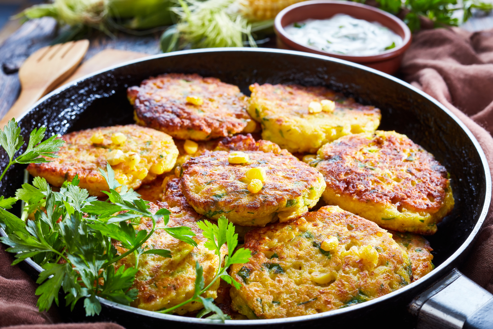 A pan of corn fritters with some greenery on the side. In the background there are cooking utensils like a wooden spatula, a bowl of sauce, and a cloth napkin