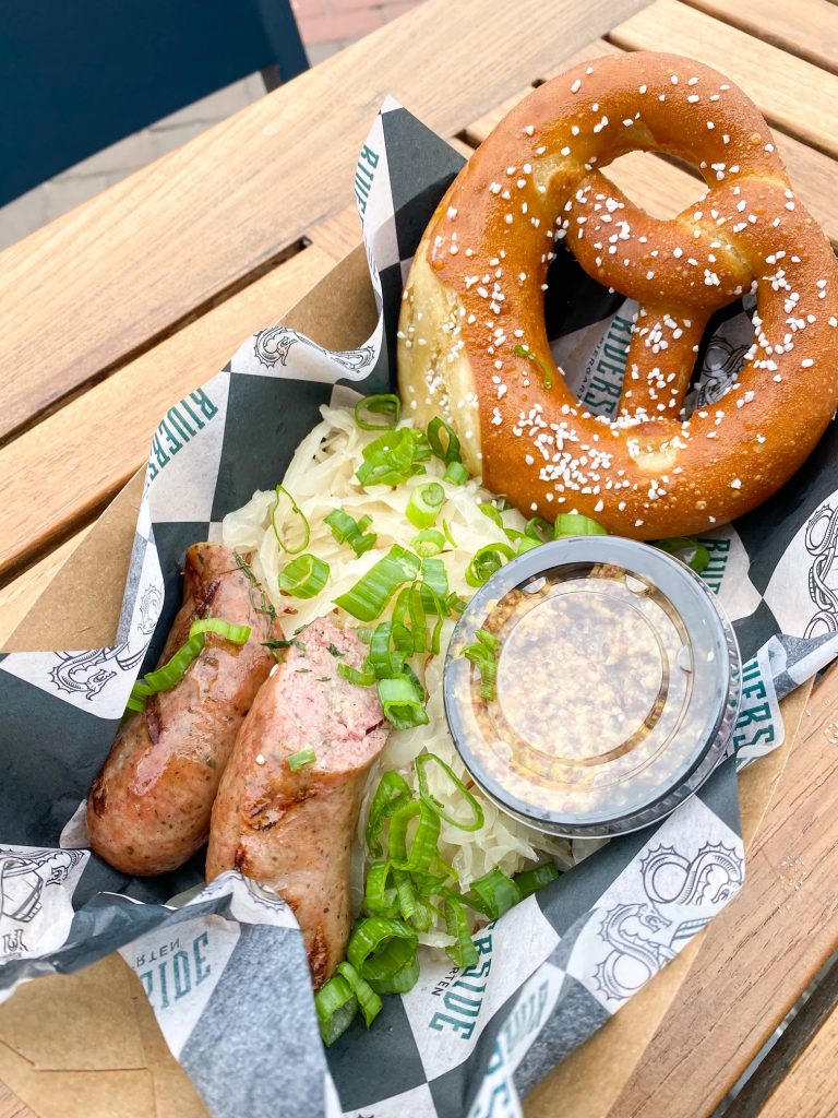 A basket of food that has sausages, sauerkraut, a cup of mustard, and a large pretzel. It is in a paper basket that has black and white checked paper in it. There are lots of scallions covering the food. The basket of food is sitting on a wooden slat table. 