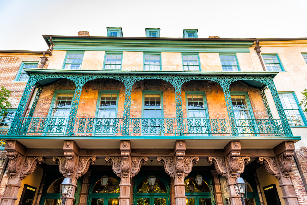 The exterior of the top of the Dock Street Theater. There is intricate iron work on the patio that is painted green. The building is brick and plaster with the plaster painted an orange color. The window trimmings are painted green. 