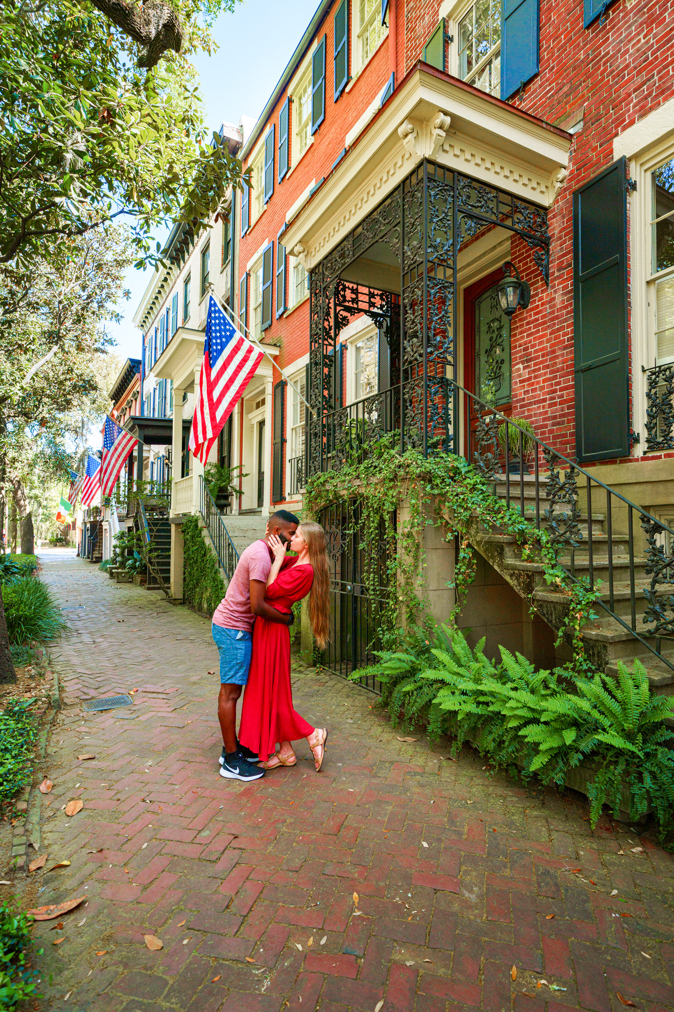 A couple standing and kissing on East Jones Street in Savannah. The street is lined with historic homes hanging American flags. It is a cobblestone sidewalk and there are trees on the other side. The house closest to the couple is brick with wrought iron stairs and overhang that is covered in vines. The woman in the couple is wearing a red dress and her long hair is down. The man in the couple is wearing a red heather shirt with denim blue shorts. 