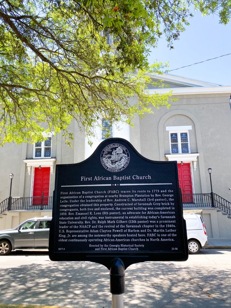 A large black plaque on the sidewalk in front of the First African Baptist Church. It details the history of the church. Behind the sign you can see a large grey building with stained glass windows with white trim and two sets of large red doors. There is a tree that is hanging over the black sign. 