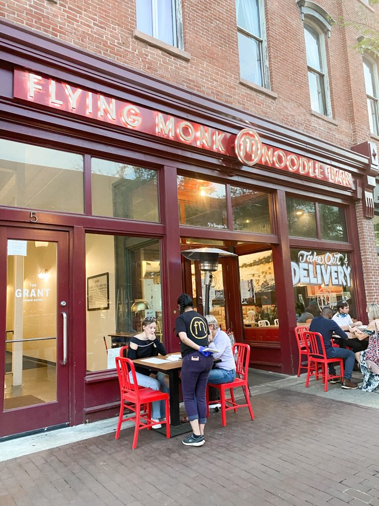 The exterior of the Flying Monk Noodle Bar in Savannah. The building is full of windows with the restaurants logo and an infographic that says 'take out and delivery'. There are people sitting outside of the restaurant and sets