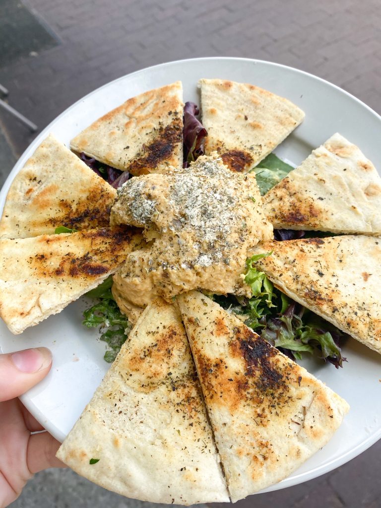 A plate of fresh hummus and pita bread on a bed of greens. It is on a white plate and someone is holding the plate. 