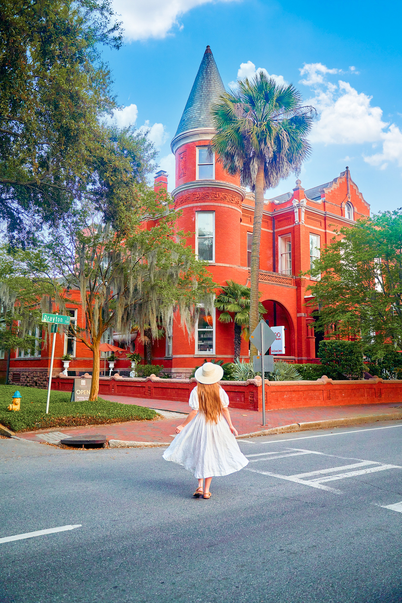 A woman with long hair wearing a white sundress and sun hat standing in the road in front of the Forsyth Mansion. Forsyth Mansion is a large brick home with turrets, lots of windows with white trim, and a courtyard. There are a lot of trees in front of it and shrubs and it is on the corner of the street. A great place to see during your 3 days in Savannah