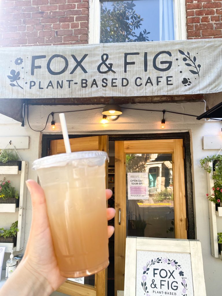 A hand holding a limeade drink with a lid and a straw in front of a store front. The store front is 'Fox & Fig Plant Based Cafe'. There is a large set of wooden French doors that lead inside the cafe. You can see some flower boxes on either side of the door with yellow, pink, and orange flowers. There is also a a standing sign that is painted white with the shops name and logo on it. 