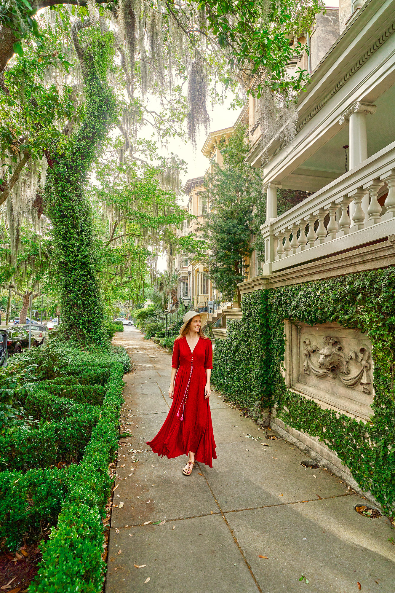 A woman in a red dress standing on the sidewalk of a beautiful street in Savannah. One side of the sidewalk is lined with historic homes covered in ivy and other greenery. The other side has trees and boxwood shrubs