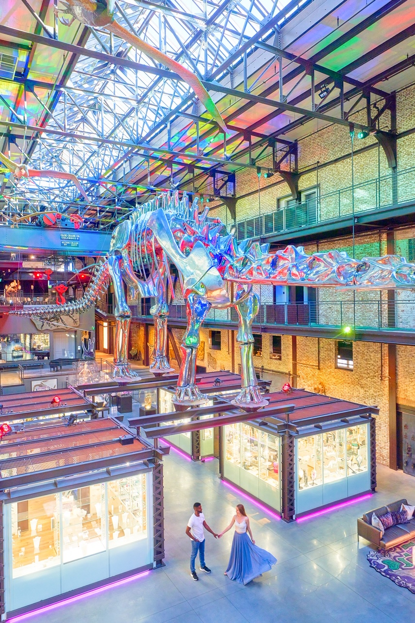 A couple standing in the lobby of the JW Marriott Hotel in Savannah. The lobby has glass cases full of artifacts, a metal dinosaur skeleton hanging from the ceiling, and its lit up with different colored lights. The woman in the couple is wearing a white top and long flowy blue skirt. The man in the photo is wearing a white shirt and blue jeans. Its a great Savannah photo spots