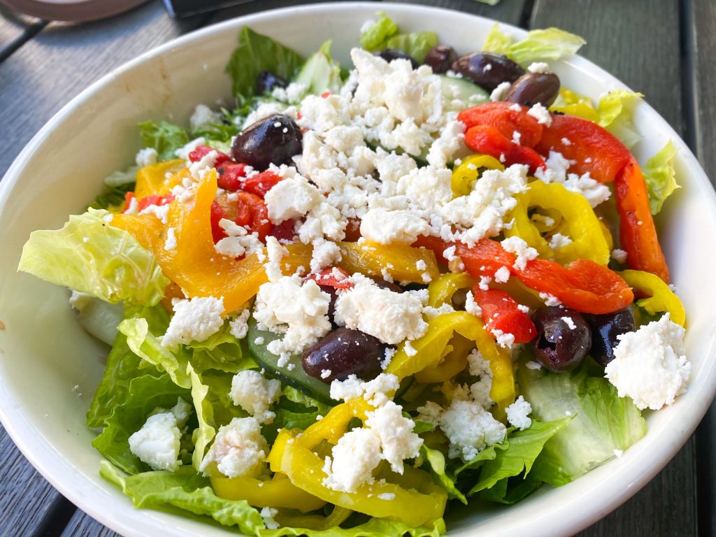 A large Greek salad full of romaine lettuce, red peppers, yellow peppers, red onion, olives, picked jalapenos and feta cheese in a white bowl. 