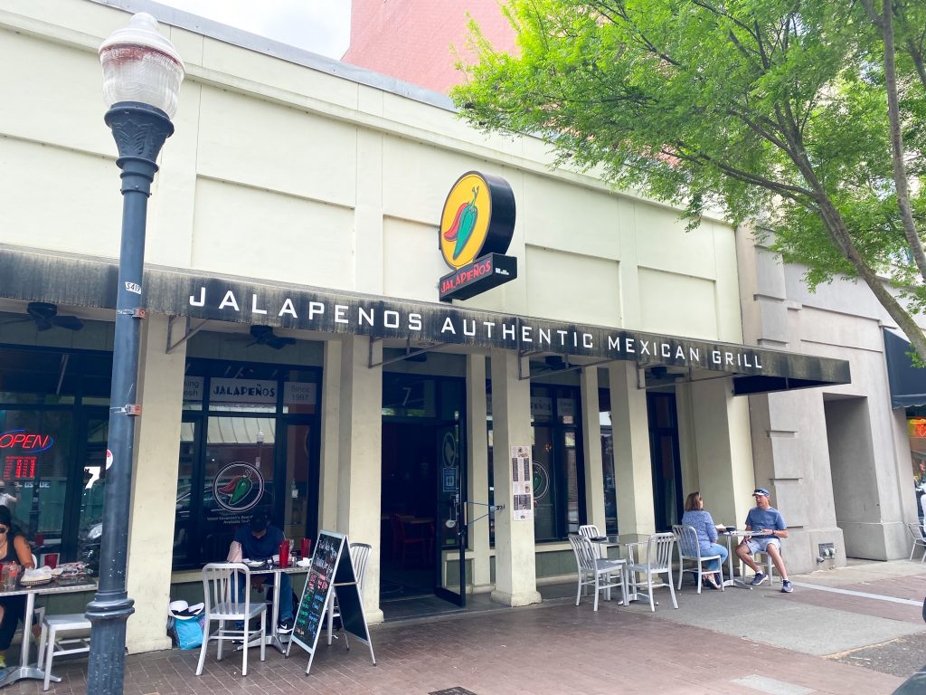 The exterior of Jalapenos Authentic Mexican Grill. It is a white building with a sign with a picture of jalapenos on it and a black over hang with the name on the restaurant. There are people sitting at aluminum tables in front of the restaurant. 