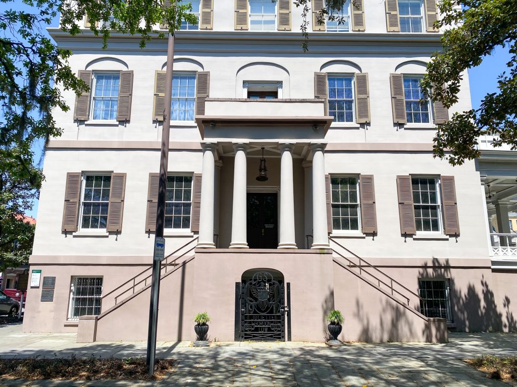 The exterior of the Juliette Gordon Low birthplace. It is a large historic building that is cream and tan with lots of windows with tan shutters. There are two stairways leading up the the large columned front porch that has the front door. It is a sunny day and there are trees and shrubs around the building. In front of the front porch is a plaque with the Girl Scout logo on it and more information on the building. 