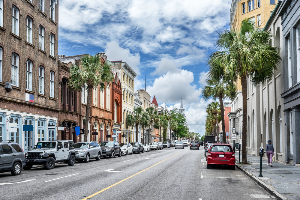 A view of King Street, the most popular shopping street in Charleston. There are lots of historic buildings that have been turned into shops, palm trees, and cars parked on the side of the street. It is a sunny day with big fluffy clouds. 