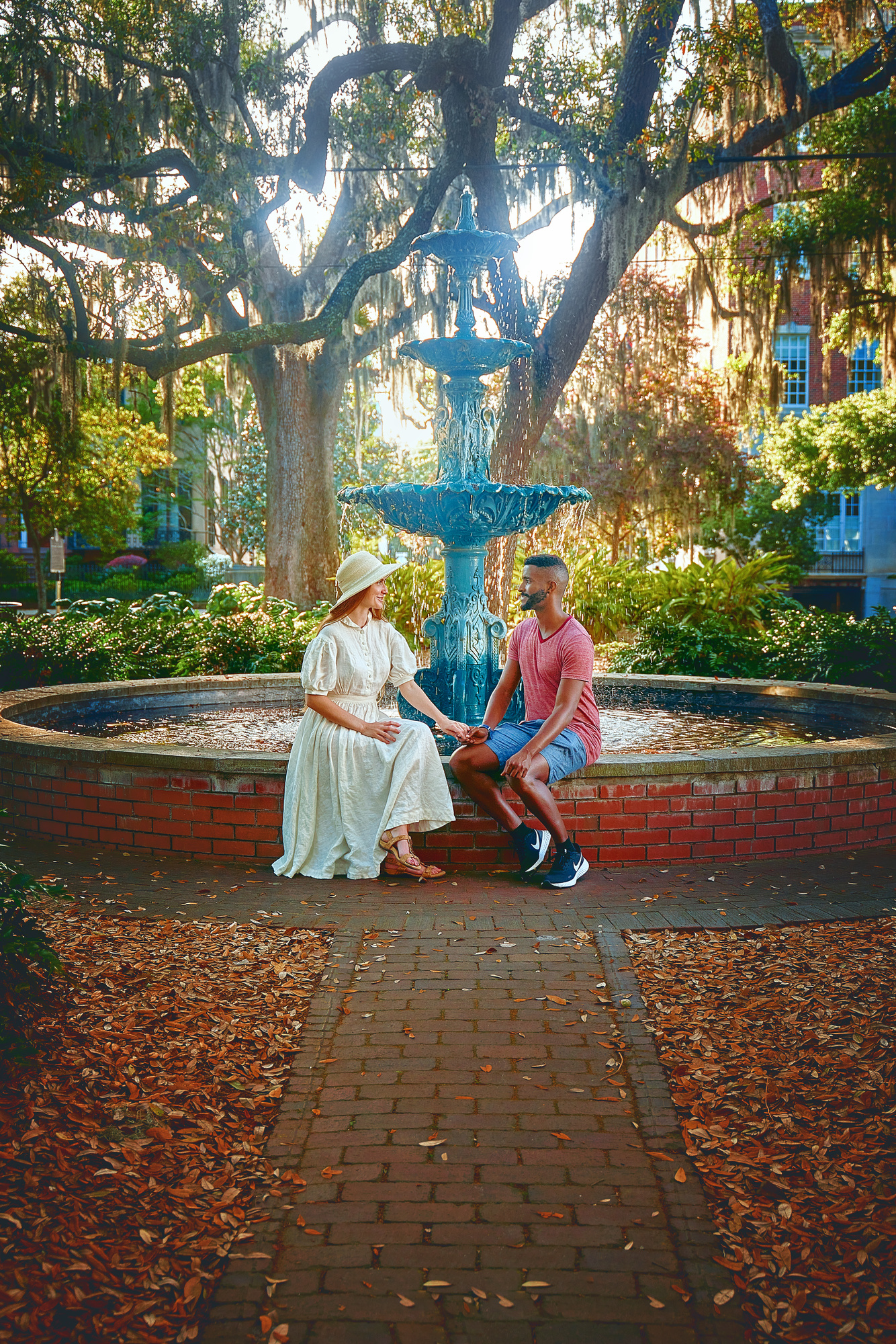 A couple sitting in front of the fountain at Lafayette Square. The Fountain is aged copper, so it is green, and the pool is made of antique brick. Behind the fountain are lots of trees with Spanish moss and shrubs. You can just barely see buildings through the trees. The woman in the couple is wearing a white dress with a white sun hat. The man in the couple is wearing a red heather shirt and denim blue shorts. They are sitting and looking at each other while holding hands. 