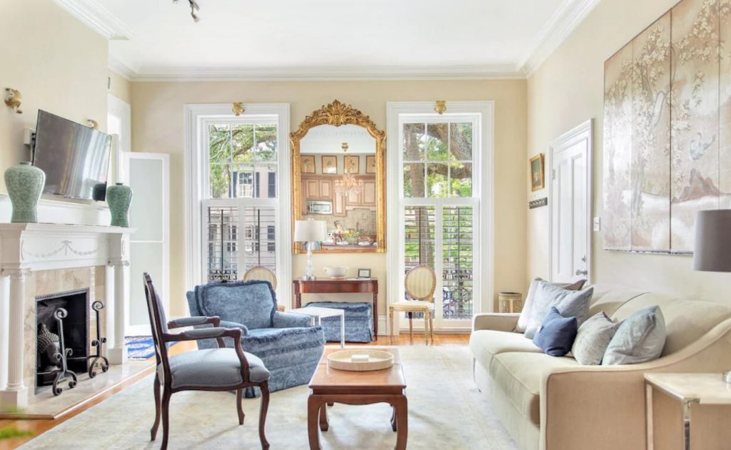 The interior of a private home in Savannah. There is a cream colored couch with different colored blue pillows, blue chairs, a fireplace, and a gold ornate frame. There are two large windows on either side of the gold frame. 