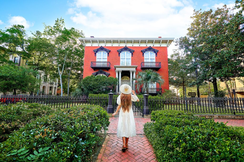 A woman standing on the sidewalk in front of the Mercer Williams House. The woman is wearing a white sundress, has long hair, and is wearing a white sunhat. She is standing on a brick sidewalk or courtyard surrounded by boxwood shrubs. The house has a black wrought iron gate, a front yard with more boxwood shrubs, and trees. The house is square with a brick façade, large arched windows with intricate black trim, and small balconies on two of the windows. 