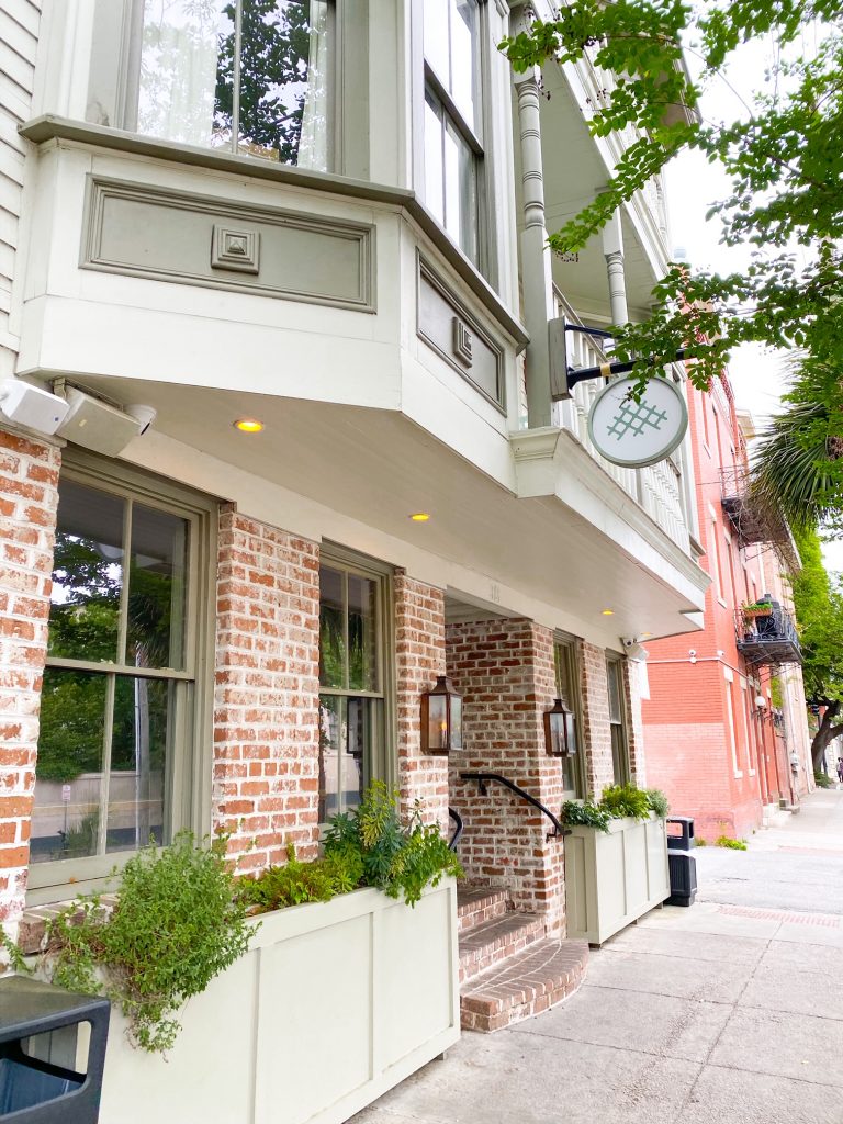 The exterior of a restaurant in Savannah. It has large pale green flower boxes full of greenery on either side of a doorway. Most of the exterior is antique brick and there are stairs leading inside the building that are also brick. Next to the stairwell are two copper sconces. The second story of the building sticks out further than the bottom and is cream and a gray-green with windows and what looks to be a porch. There is a sign hanging from the second story with a symbol that looks like a cross stitch pattern. A great spot on your 3 days in Savannah