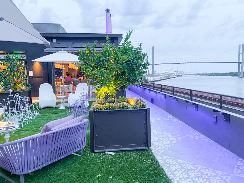 The Myrtle and Rose Roof Top Bar in the New Plant Riverside District of Savannah. It has a view of the river and is decorated in purple and white. You can see the walls and floors of the roof are painted purple, there is a purple bench, and white seats under a white umbrella. There is also fake grass on the roof top where there is a wire metal white and purple chess set. On the rooftop there are planter boxes with small plants and shrubs. You can see the entrance to the indoor bar area at the end of the rooftop. A fun way to end one of your 3 days in Savannah