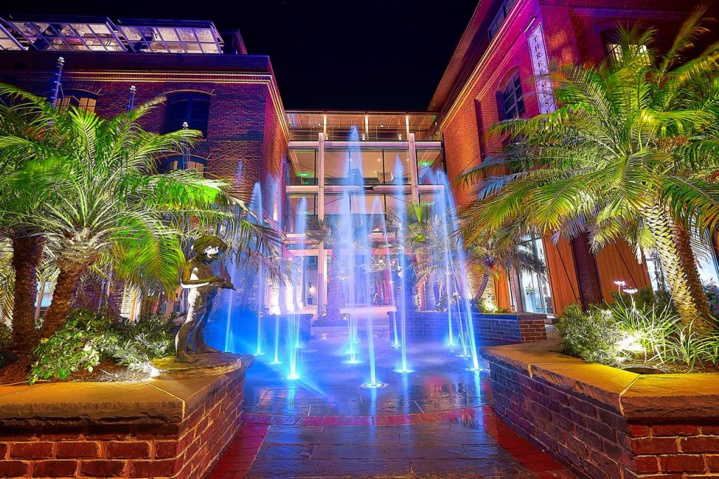 A water feature in the sidewalk in front of a large building in front of the New Plant Riverside District in Savannah. It is night time and the water feature is fountains that shoot up from the sidewalk and have colors around them. There are two raised plant beds that have greenery, palm trees, and sculptures in them. The building behind the water feature is made of brick and looks like a hotel. 