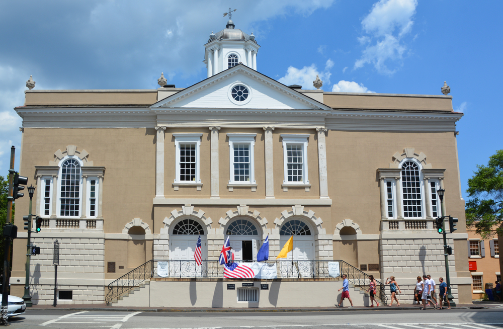 The exterior of the Old Exchange and Provost Dungeon in historic South Carolina. The building is tan and a colonial style with flags on the steps in front of it. 