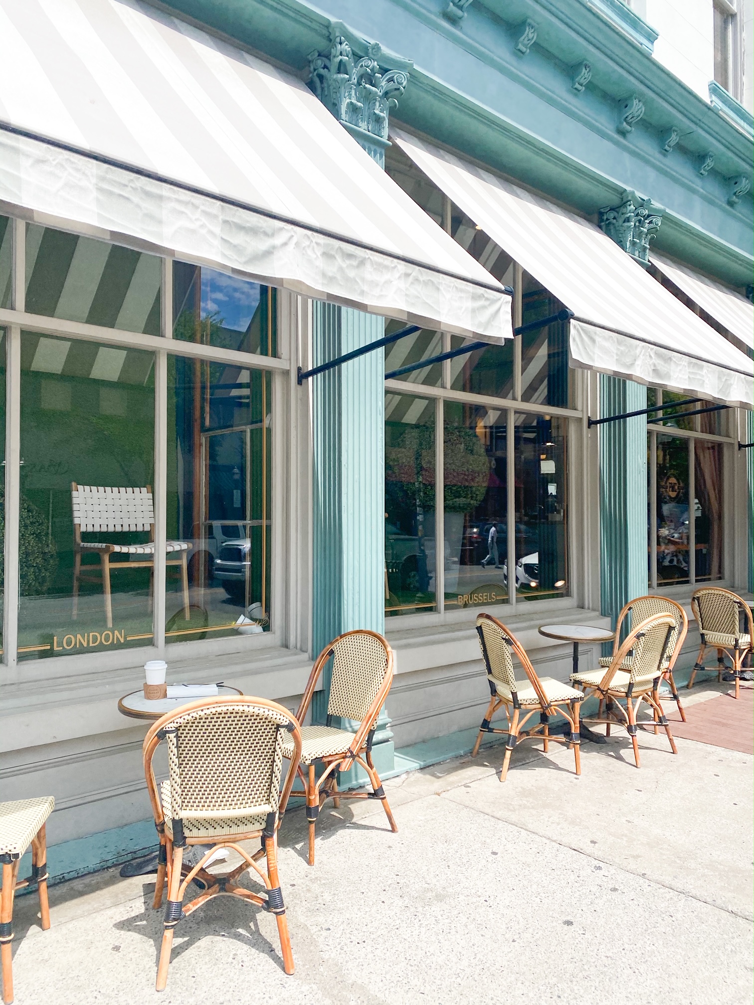 The exterior of the Paris Market in Historic Savannah Georgia. The building is painted a pale blue green and has cream and greyish tan striped awnings above the large glass windows. There are sets of outdoor dining tables and chairs that  are wooden with woven backs and seats. 