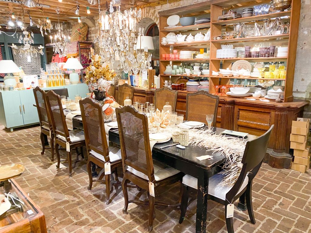 The interior of the Paris Market in Savannah Georgia. The main space is a long antique table with antique chairs. The table is elaborately decorated with glassware, a table runner, and dishes. In the background are cabinets full of dishware of all colors, trinkets, and lamps. There is also large glass chandeliers hanging from the ceiling and the floors of the store are brick. 