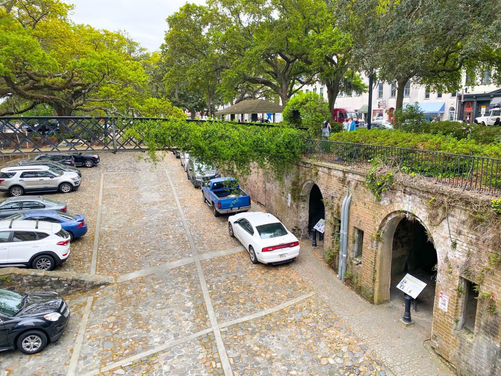 An old sunken parking lot in the historic district of Savannah Georgia. It looks very old with cobblestone and no real parking spot lines. There are cars neatly parked in the middle and on the side. On the side, there are two arch ways leading to what looks like a tunnel. Each archway has an infographic stand in it. There is grass and greenery over the tunnel and over a wrought iron bridge that crosses the sunken parking lot. 