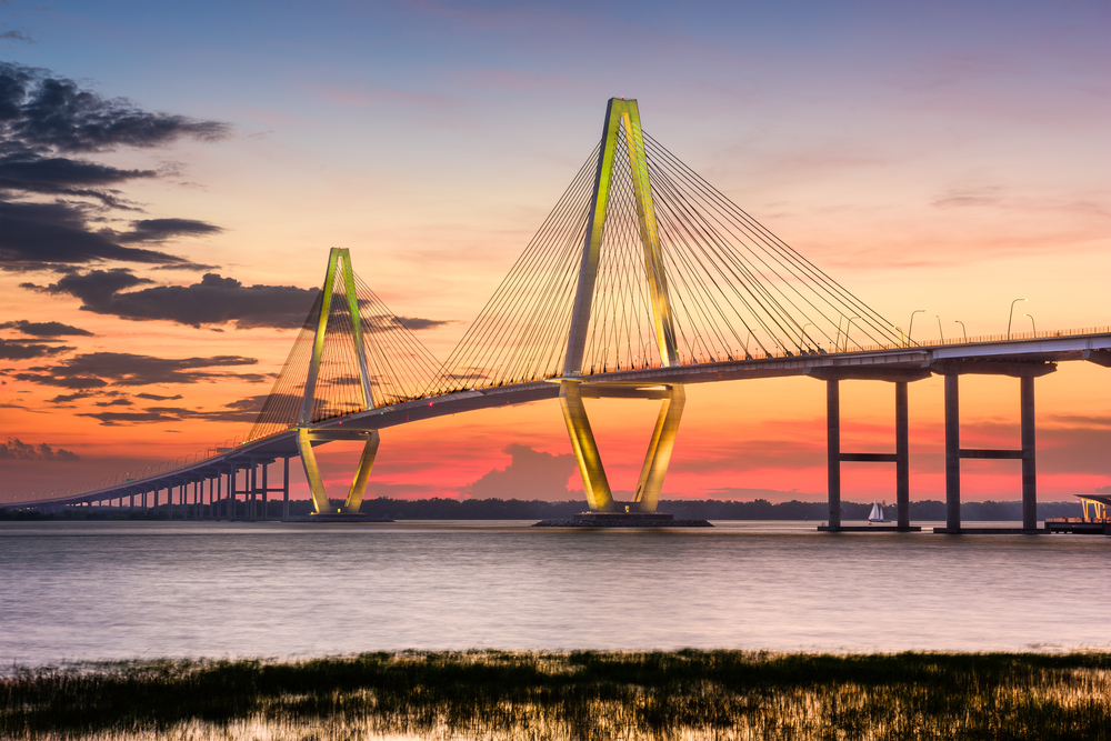 The Ravenel Bridge in South Carolina at sunset. It is a large bridge with two triangle shaped towers that have wires coming from them down onto the bridge. 